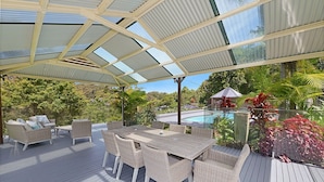 An outdoor deck with a glass roof, wicker furniture, and a pool in the background. - The Acreage Boutique Guesthouse (Picketts Valley)