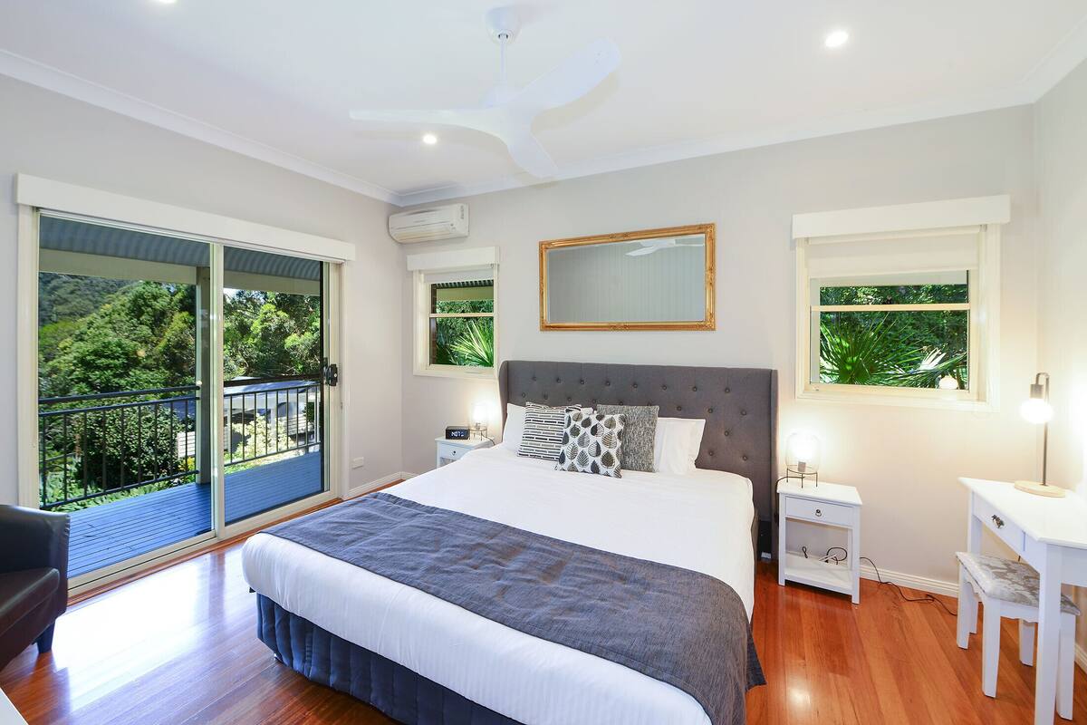 A bedroom with a large bed, a nightstand, a desk, and a chair. There is a balcony with a view of greenery.