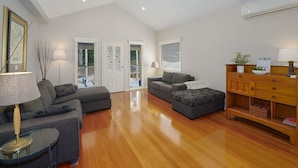 A spacious living room with a wooden floor, grey sofas, a glass coffee table, and a wooden cabinet with a plant. - The Acreage Boutique Guesthouse (Picketts Valley)