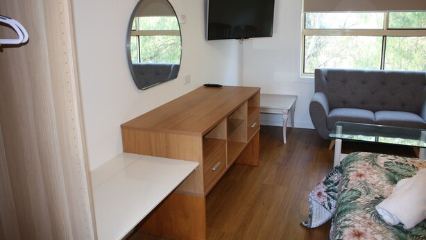A room with a wooden desk, a television mounted on the wall, a sofa, and a bed with a floral bedspread. - Coburg Motor Inn (Coburg North)