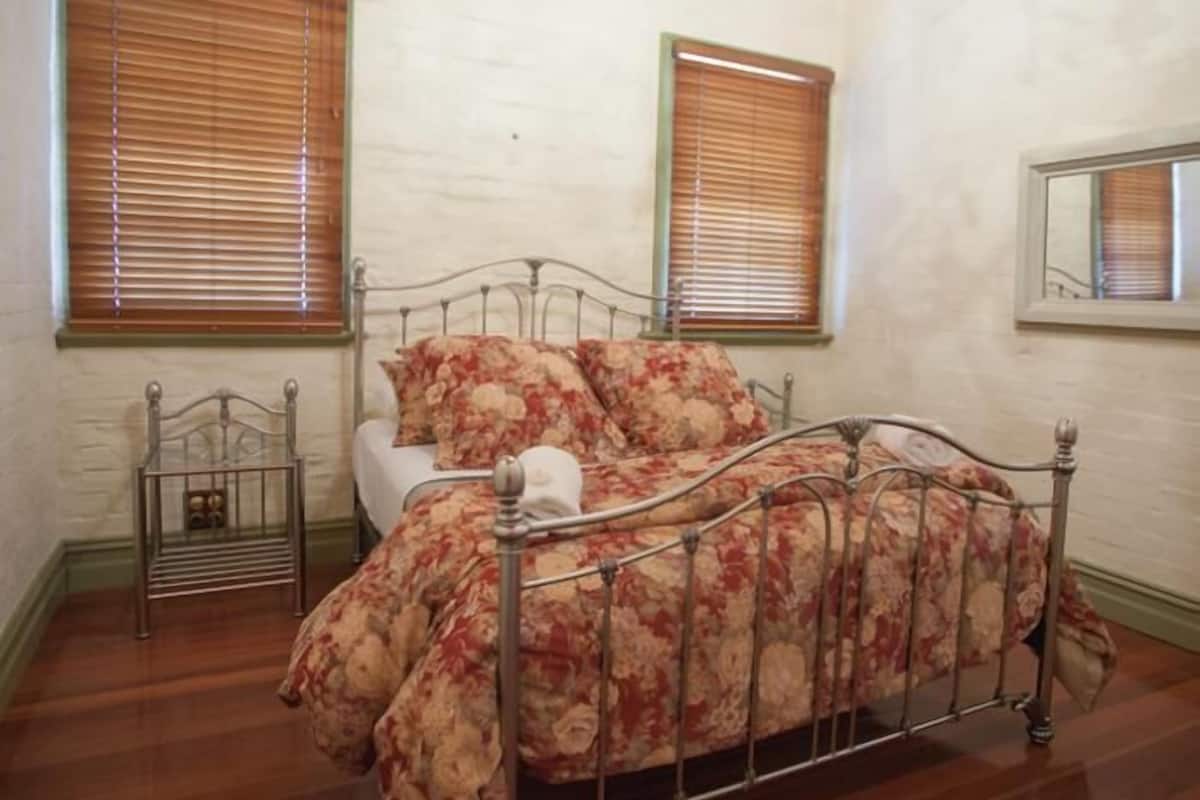 A bedroom with a metal bed frame, floral bedding, wooden blinds, and a small bedside table.