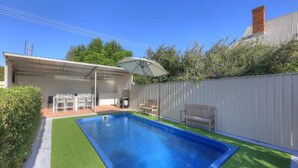 Una piscina rectangular con revestimiento azul, rodeada de césped artificial verde. Se observa un área de terraza cubierta con mobiliario exterior y una sombrilla blanca.