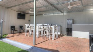 Un espacio de patio al aire libre con sillas y mesas de metal blanco, un refrigerador y un ventilador de techo.