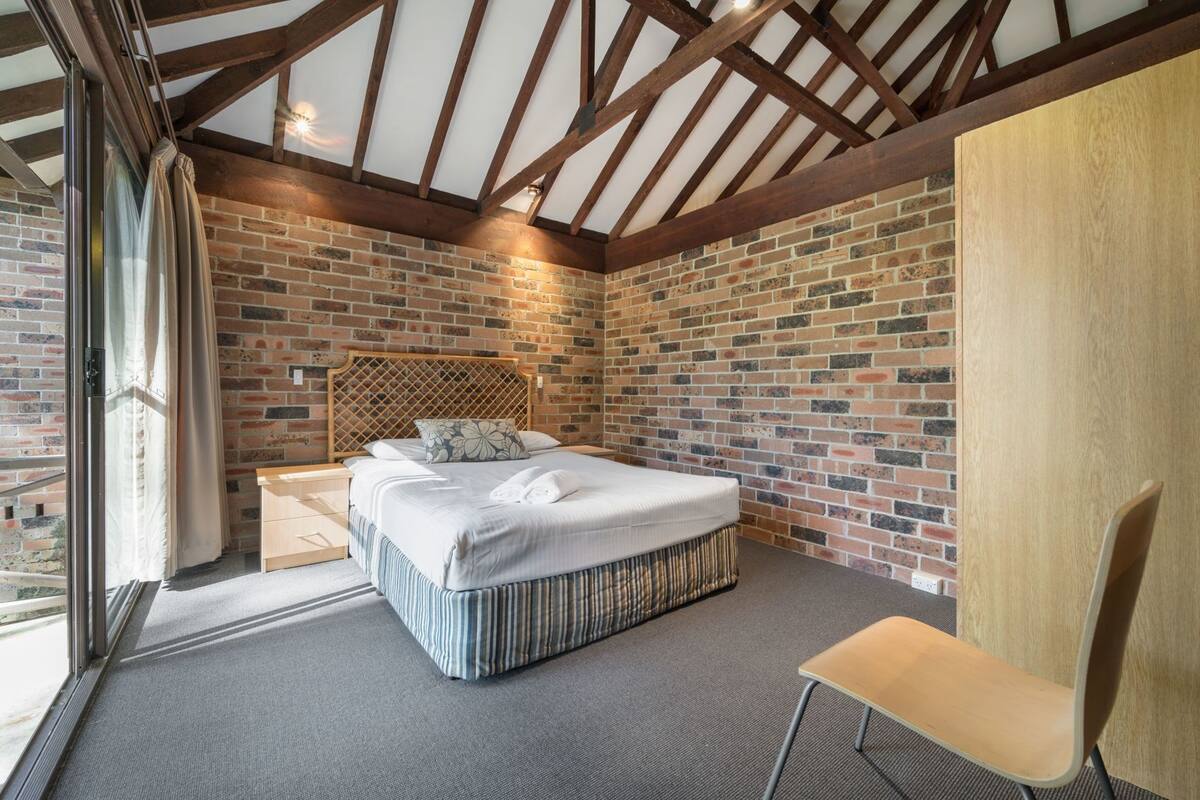 A bedroom with a brick wall, a wooden headboard, a bed with white linens, a chair, and a large window.