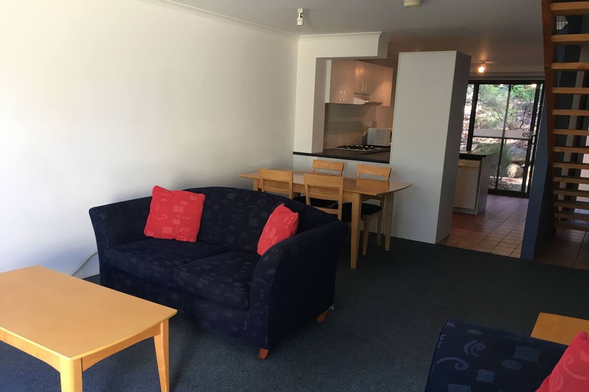 A living room with a dark blue sofa, red cushions, a wooden table, and a kitchen area in the background.