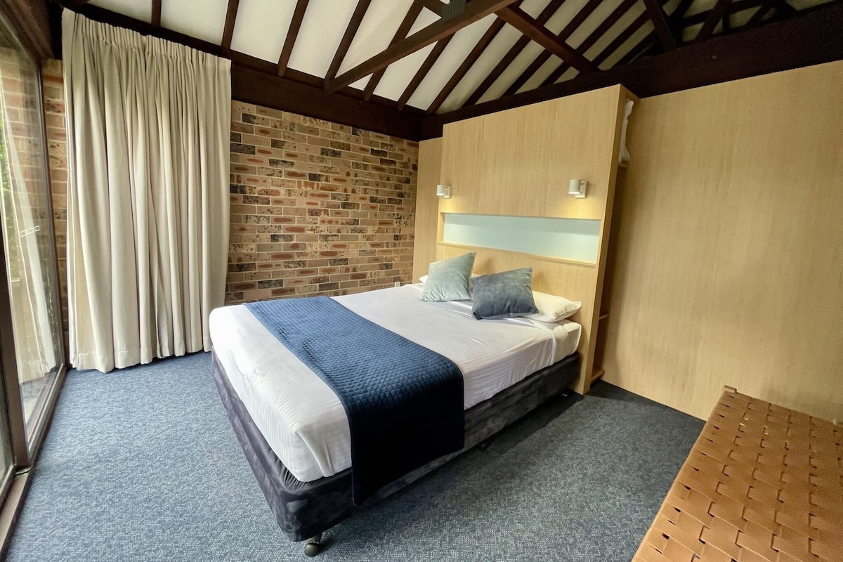 A bedroom with a bed, a brick wall, a wooden ceiling, and a window with curtains.