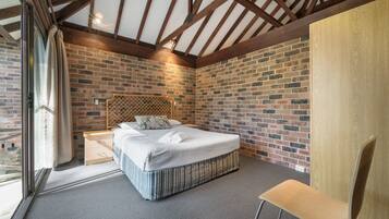 A bedroom with a brick wall, a wooden ceiling, a bed with a striped bed skirt, a wooden nightstand, and a chair.