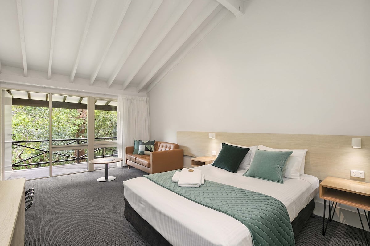 A hotel room with a large bed, a sitting area with a sofa and armchair, a small table, and a balcony with a view of greenery.