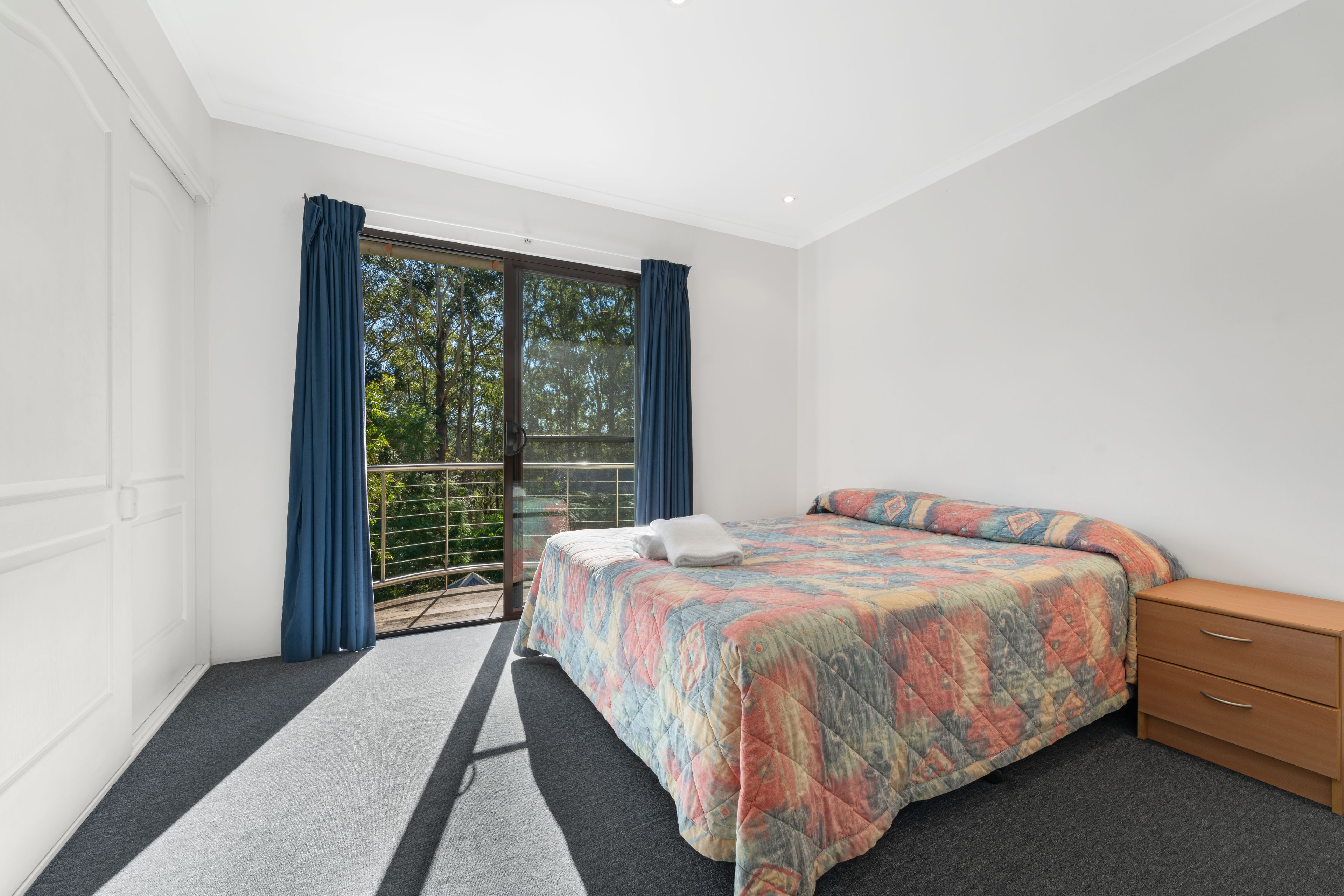 A bedroom with a bed, a wooden nightstand, a window with curtains, and a balcony with a view of trees.
