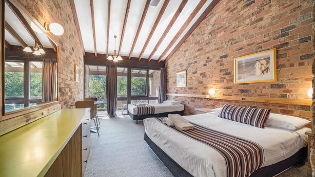 A bedroom with a large bed, a desk, and a chair. There is a brick wall and a window with curtains. - Avoca Beach Hotel (Avoca Beach)