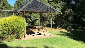 Un pabellón de madera con techo de lámina corrugada, una mesa de picnic y una base de concreto, rodeado de césped y arbustos bien cuidados.