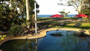 Una piscina de borde curvo, rodeada de sillones de playa y sombrillas rojas, con vista al océano al fondo.