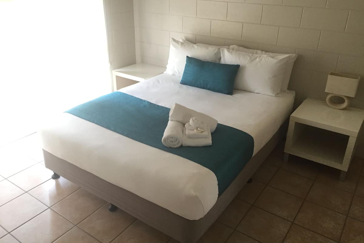 A neatly made bed with a blue accent pillow, white bedding, and a rolled towel on the footboard. There are two bedside tables, one with a lamp and the other with a small shelf. The room has a tiled floor and white brick-patterned walls.