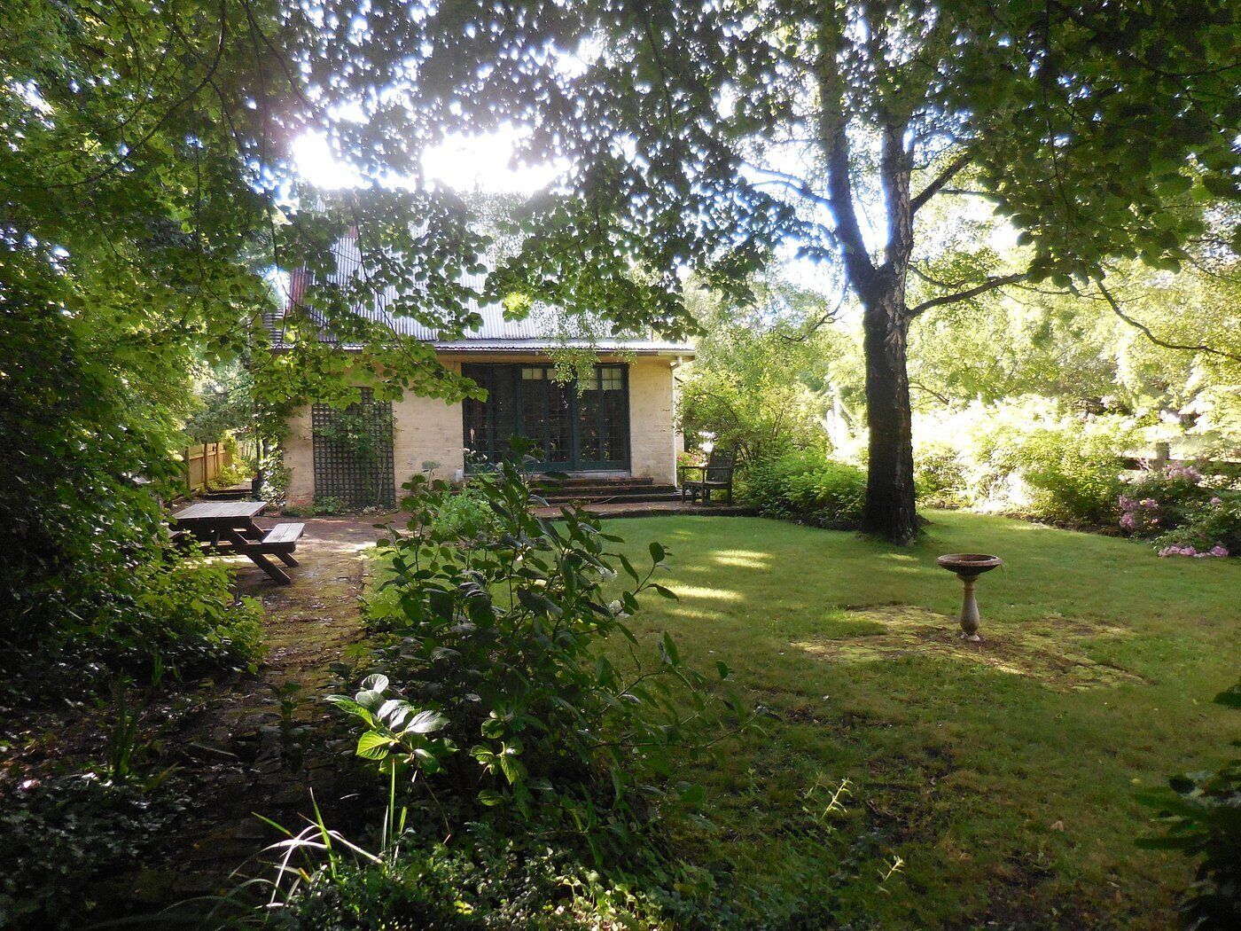 Ein kleines Haus mit Garten, einer Terrasse mit Tisch und Stühlen sowie einem großen Baum im Hintergrund.