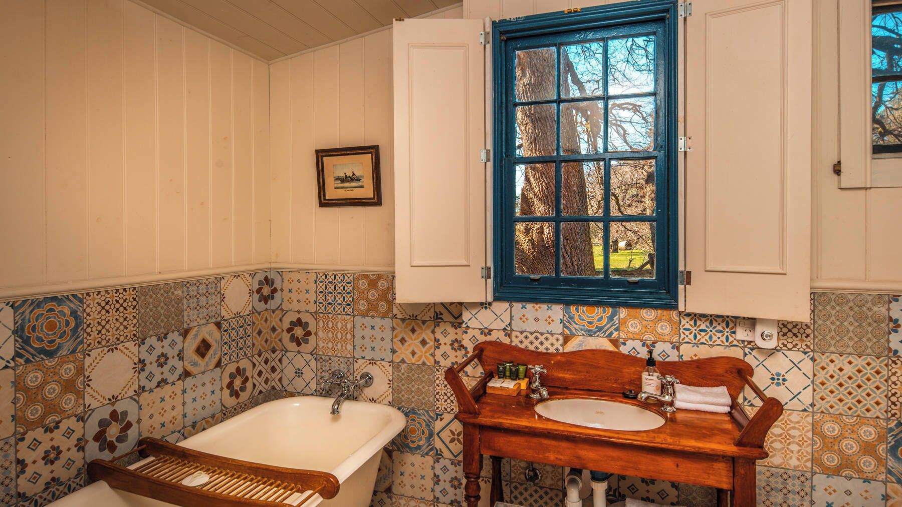 A bathroom with a bathtub, a wooden vanity with a sink, a window with blue shutters, and patterned tile walls.
