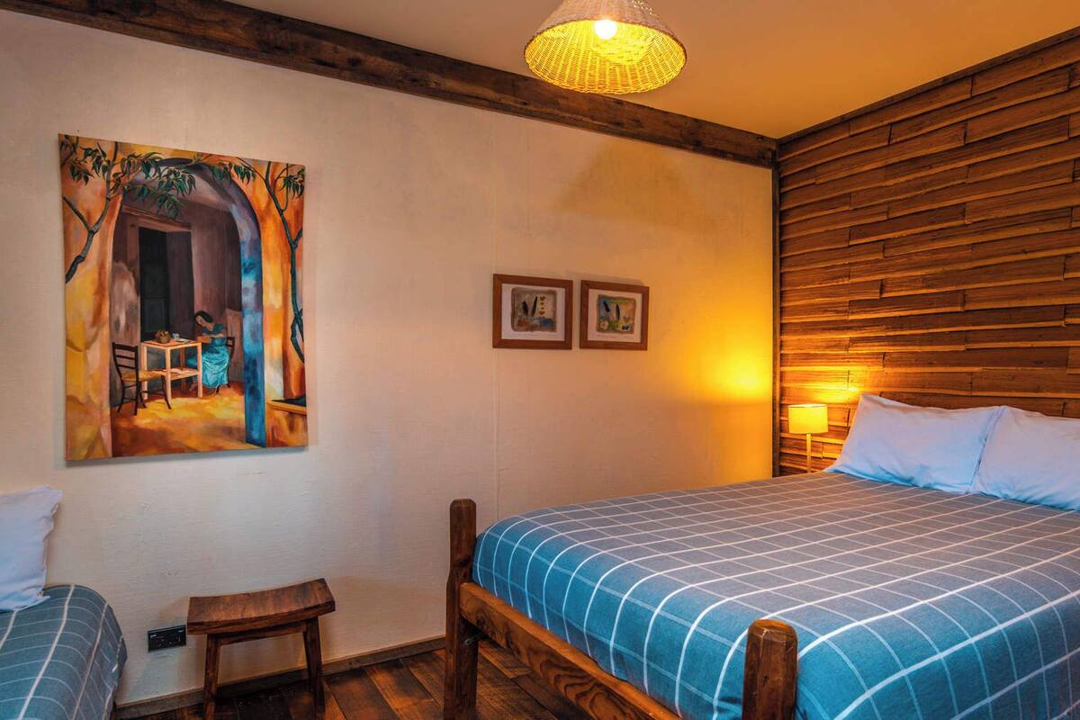 A bedroom with a wooden headboard, a bed with a blue checkered bedspread, a nightstand with a lamp, a small wooden stool, and two framed pictures on the wall.