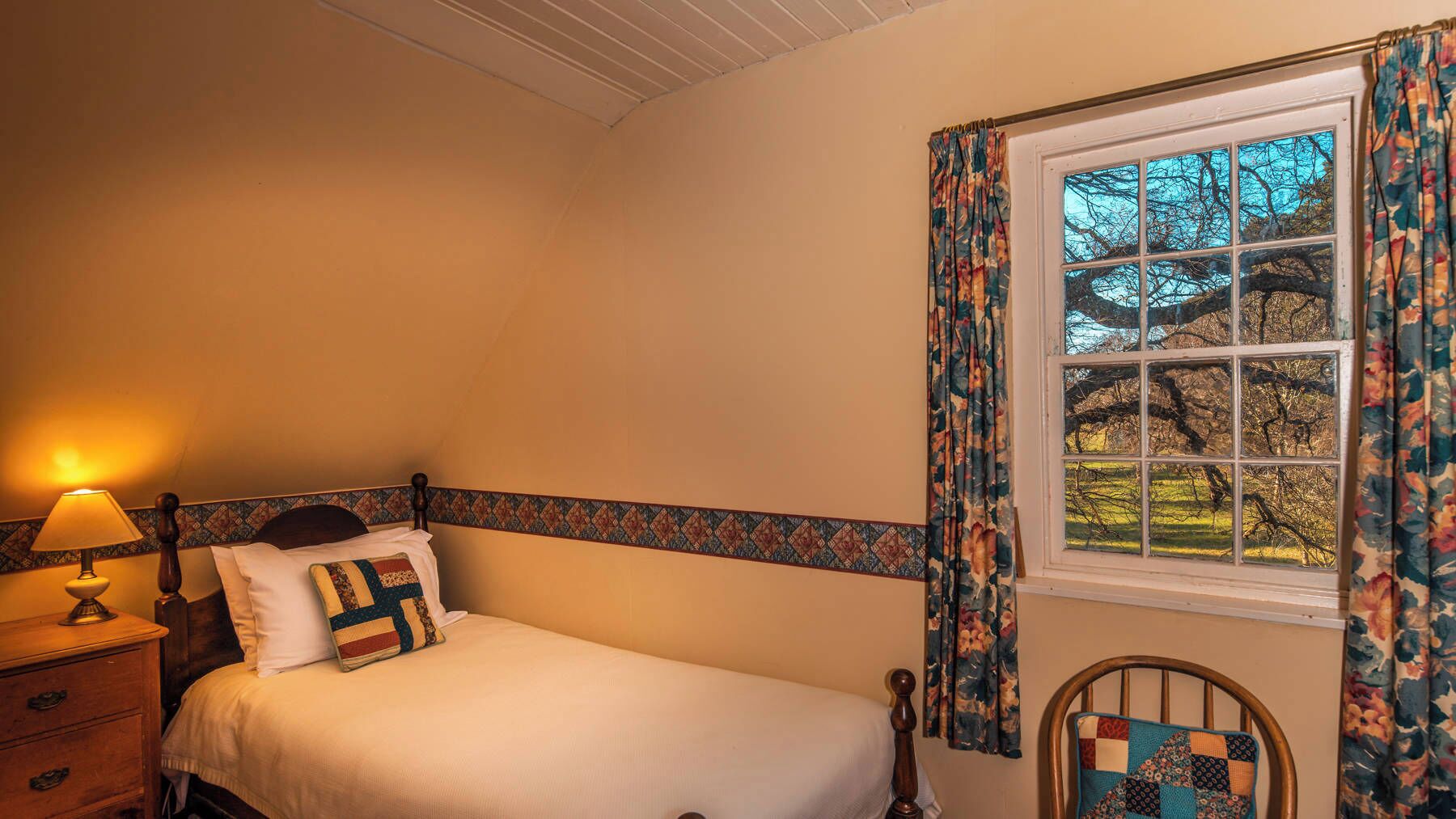 A bedroom with a bed, a nightstand, a lamp, a window with curtains, and a chair.
