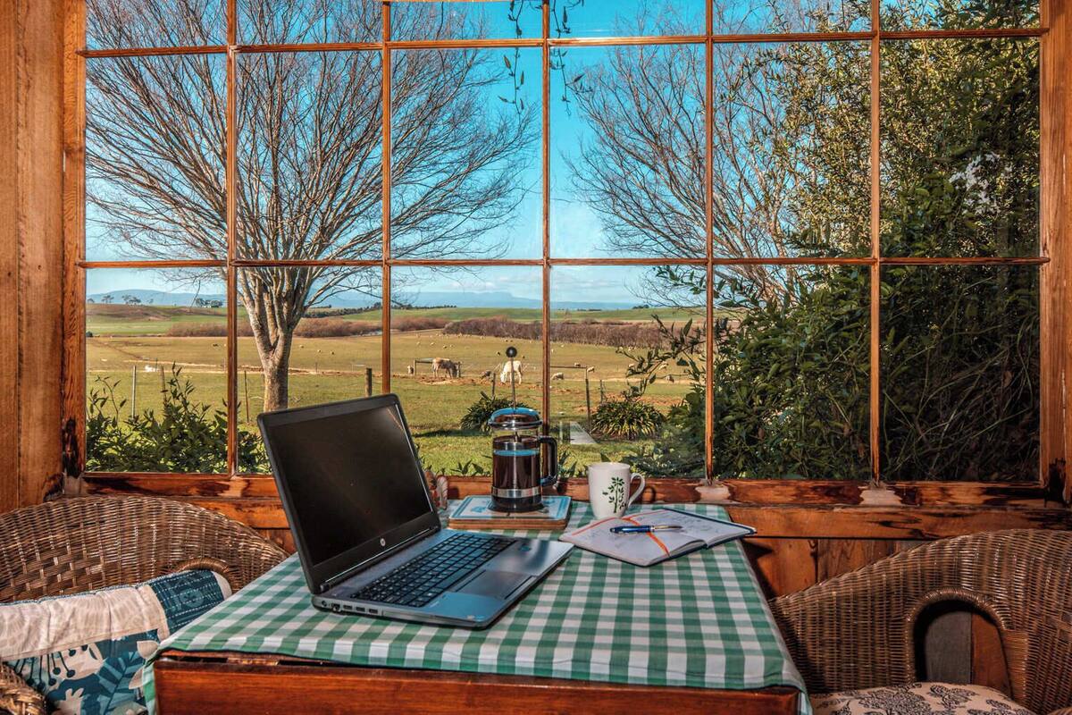 A rustic cabin with a large window overlooking a rural landscape. Inside, there's a laptop on a checkered tablecloth, a coffee pot, and a mug.