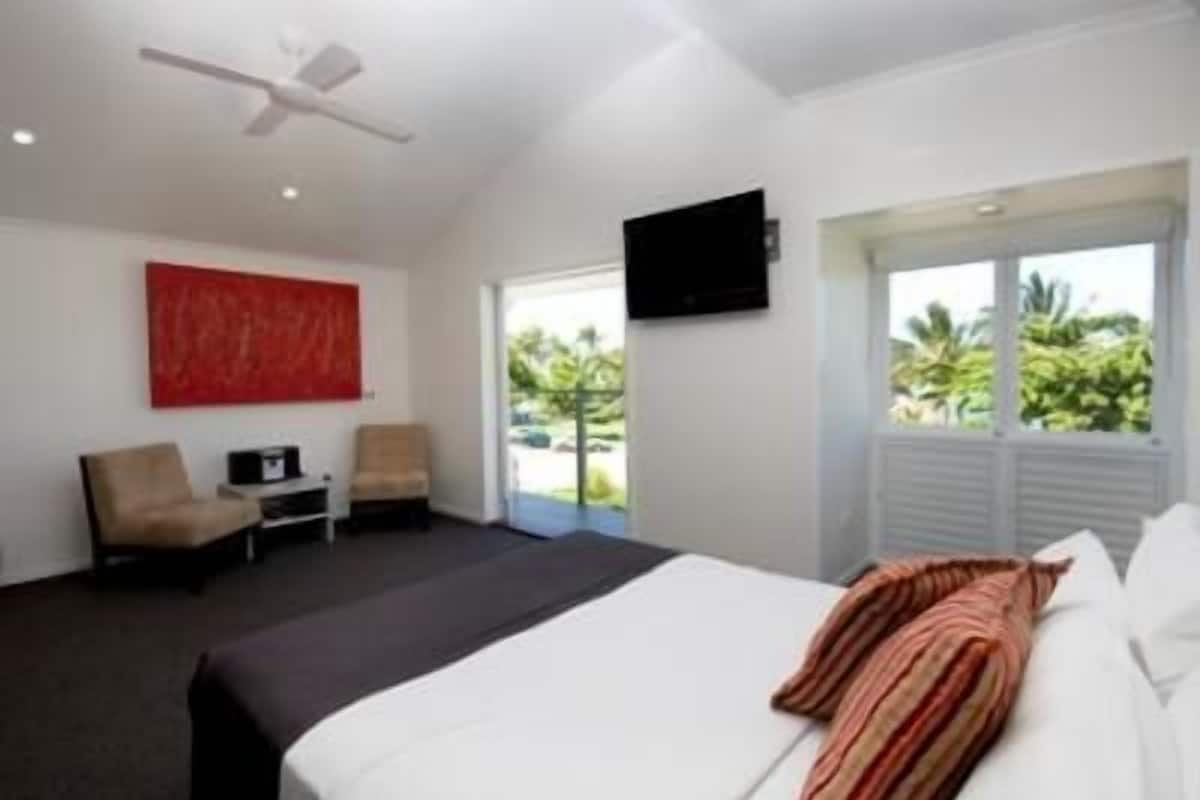 A modern hotel room with a large bed, a ceiling fan, a TV mounted on the wall, and a view of the outdoors through a large window.