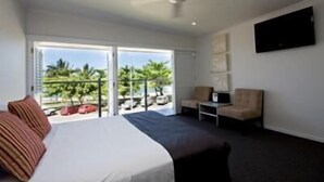 Habitación de hotel con cama, una silla, televisor montado en la pared y balcón con vistas a palmeras y coches aparcados.