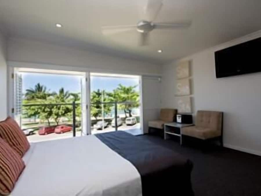 Habitación de hotel con cama, una silla, televisor montado en la pared y balcón con vistas a palmeras y coches aparcados.