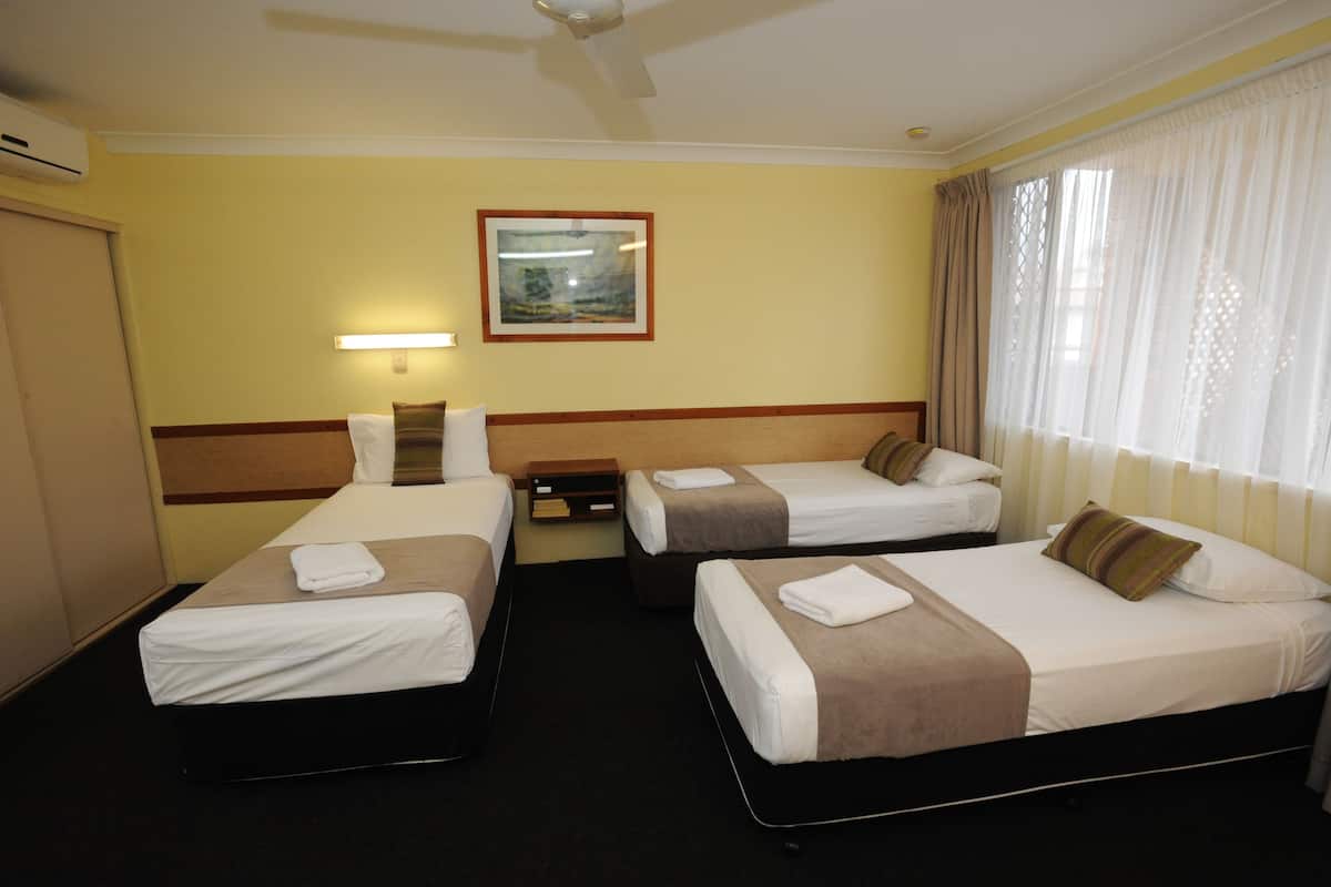 A hotel room with three single beds, a bedside table, a nightstand, a framed picture on the wall, and a window with curtains.