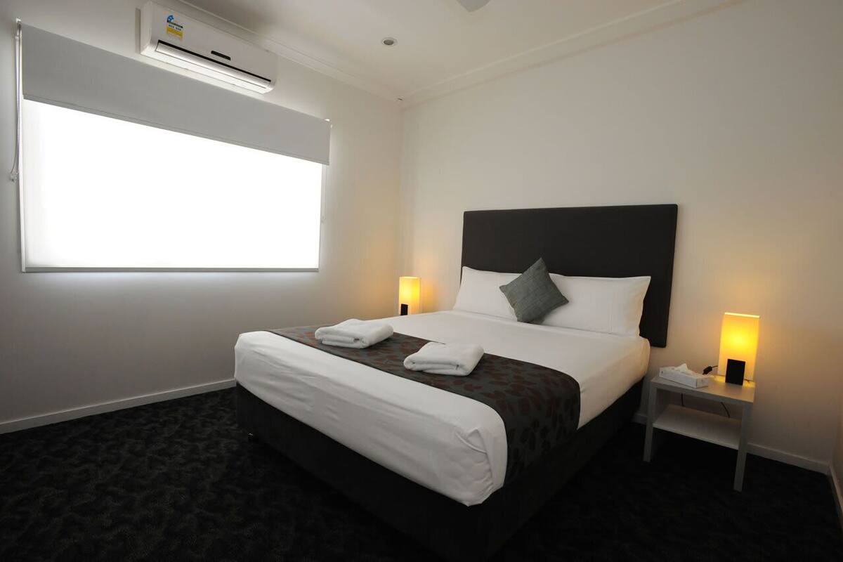 A hotel room with a large bed, two bedside tables with lamps, an air conditioning unit, and a window.