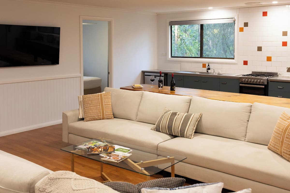 A modern living room with a sofa, a coffee table, and a TV. There is a kitchen area with a window and a tiled backsplash.