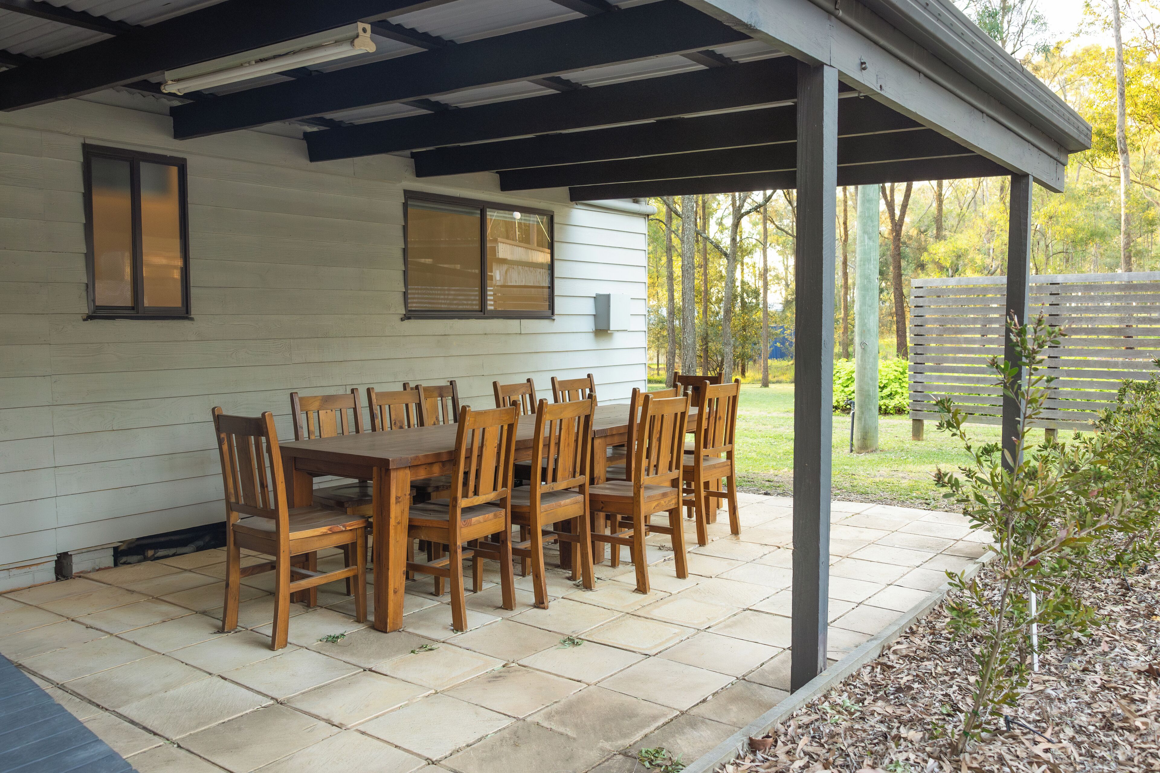 A covered patio with a wooden table and chairs, a window with blinds, and a view of the yard.