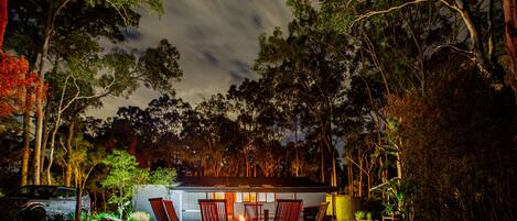 A cozy outdoor seating area with a fire pit, surrounded by trees and a small building.