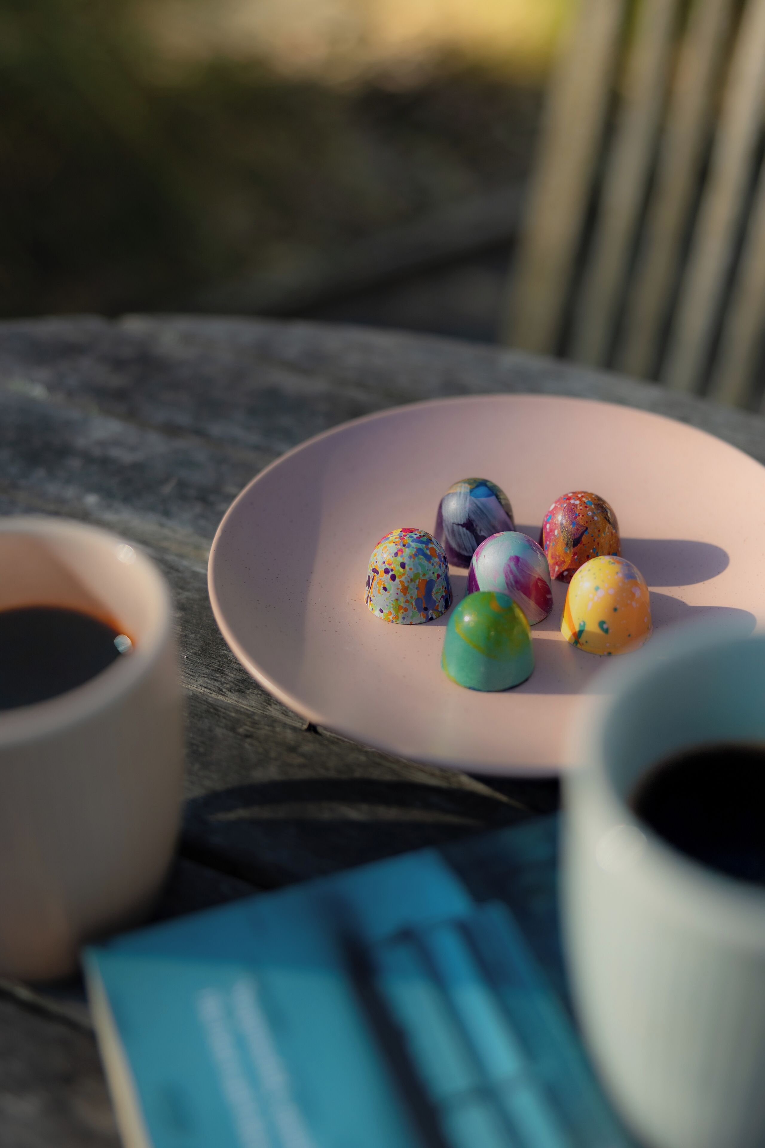 Una mesa con un plato rosa que sostiene huevos marmolados de colores, dos tazas de café y un folleto azul.