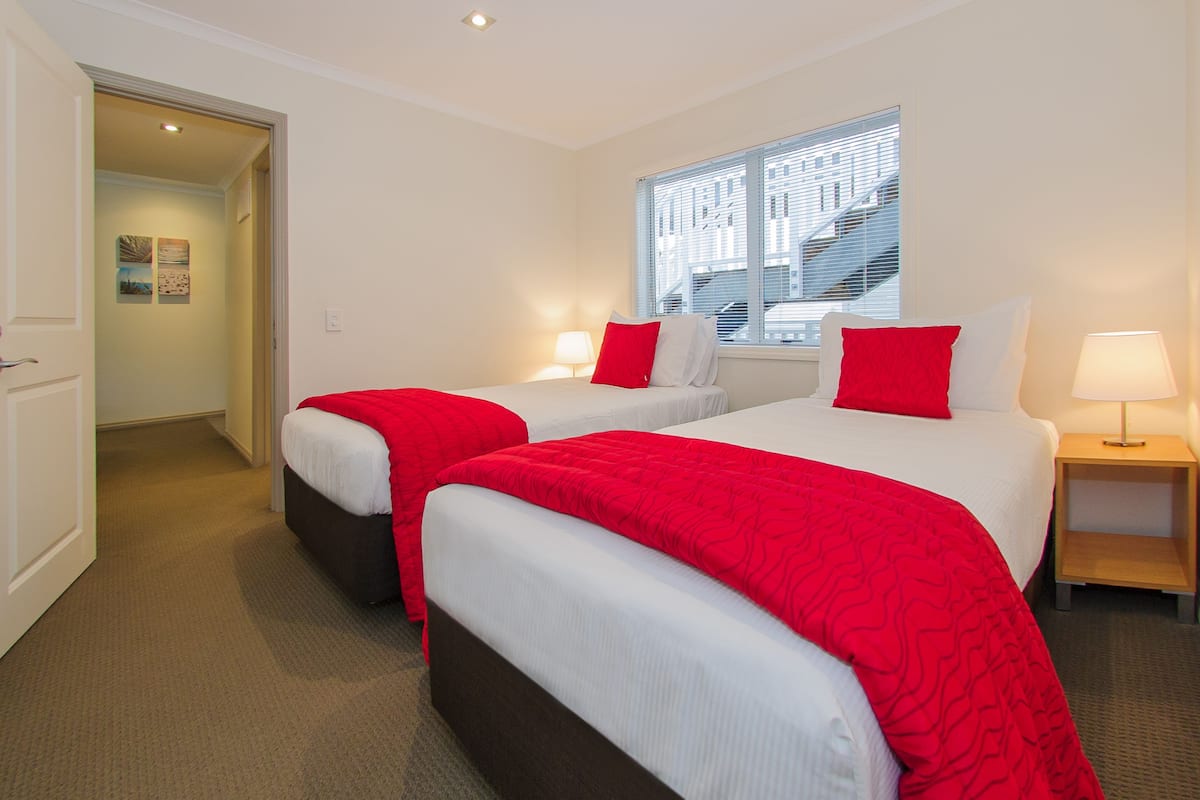 Habitación de hotel con dos camas, cada una con una colcha roja y almohadas blancas, una ventana con persianas, una mesita de noche con lámpara y una puerta que da a un pasillo.