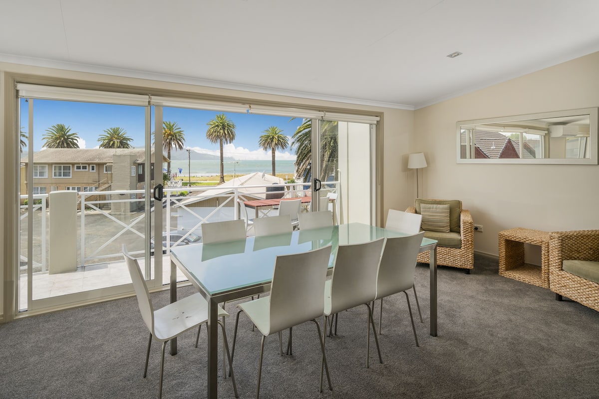 Una habitación amplia con un gran ventanal que da a un balcón, una mesa de comedor con sillas, un sofá y una pequeña mesita auxiliar.