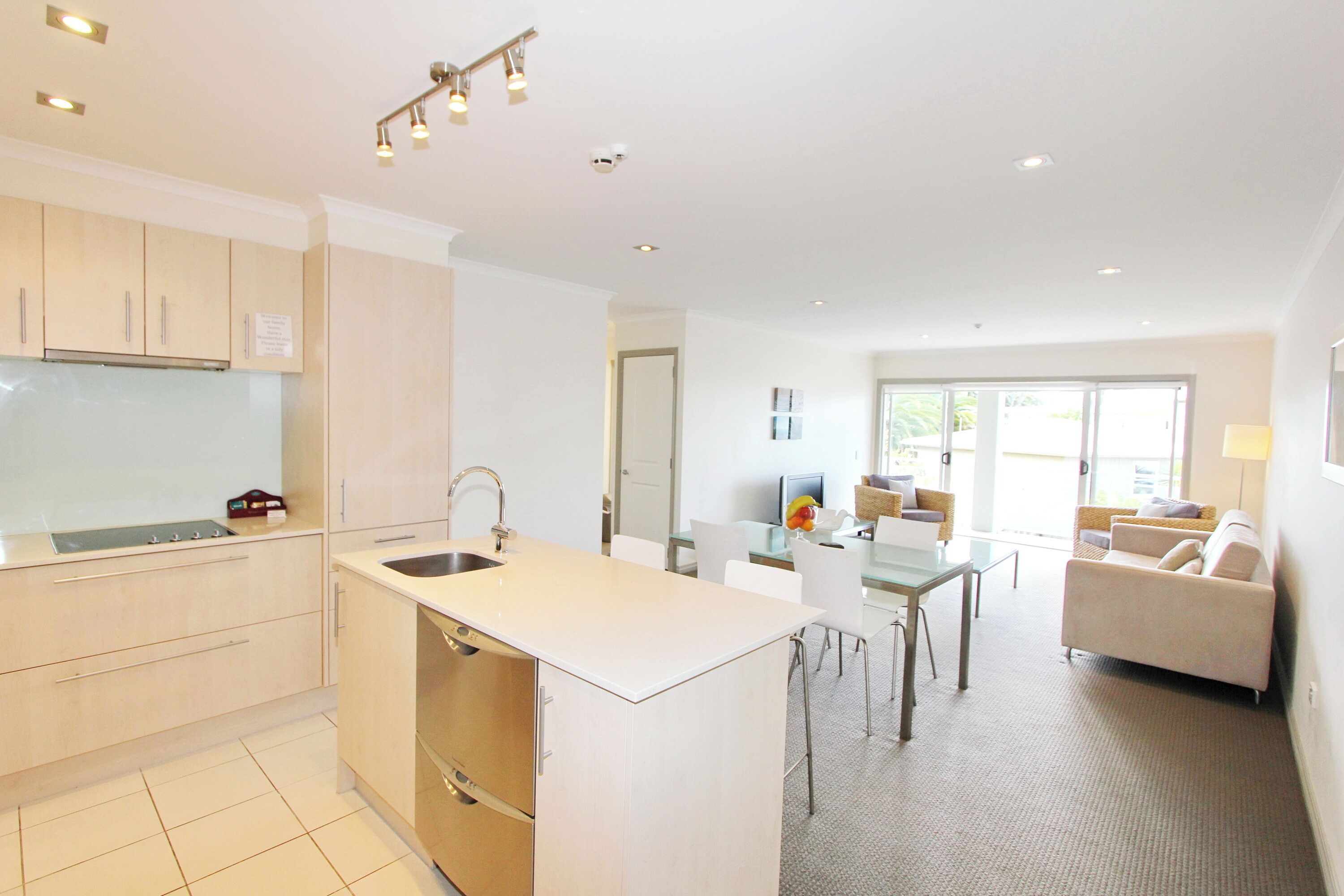 A modern kitchen with a central island, a dining area with a glass table, and a living room with a sofa and a TV.
