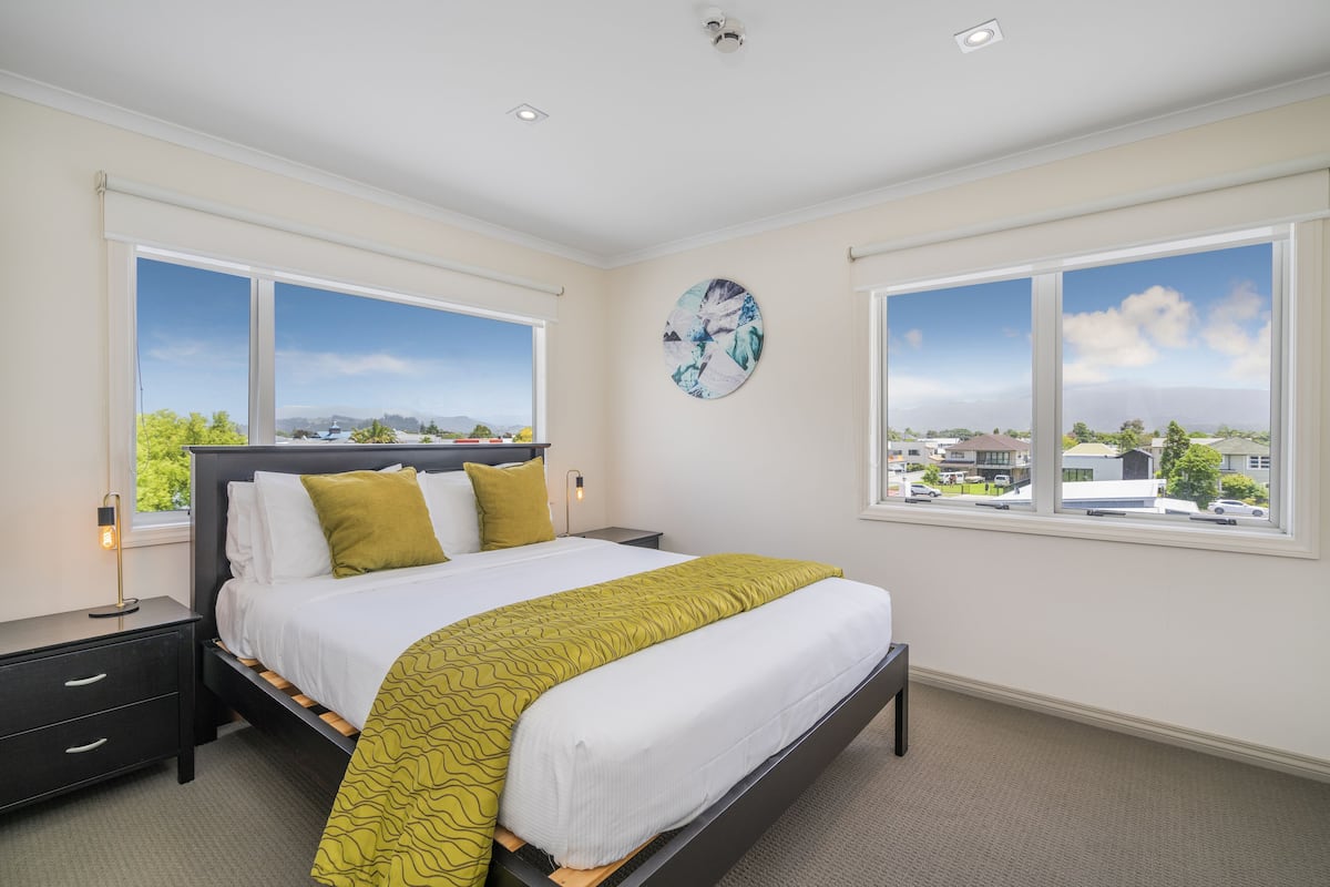 Un dormitorio con cama, mesitas de noche, un reloj de pared y ventanas amplias que dan a vistas de la ciudad.