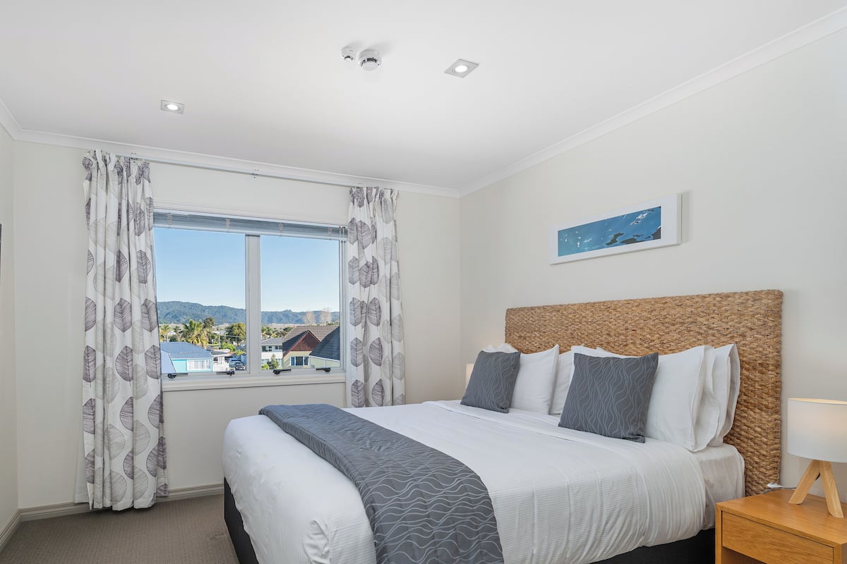 Una habitación de hotel con una cama, una mesita de noche, una lámpara, una ventana con cortinas y un cuadro en la pared.