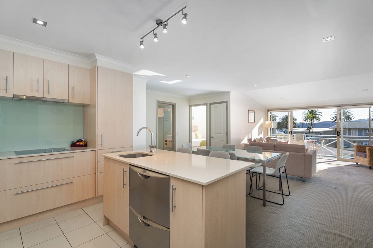 Una cocina moderna con una isla central, un comedor con mesa y sillas de vidrio, y vistas a palmeras a través de ventanas amplias.
