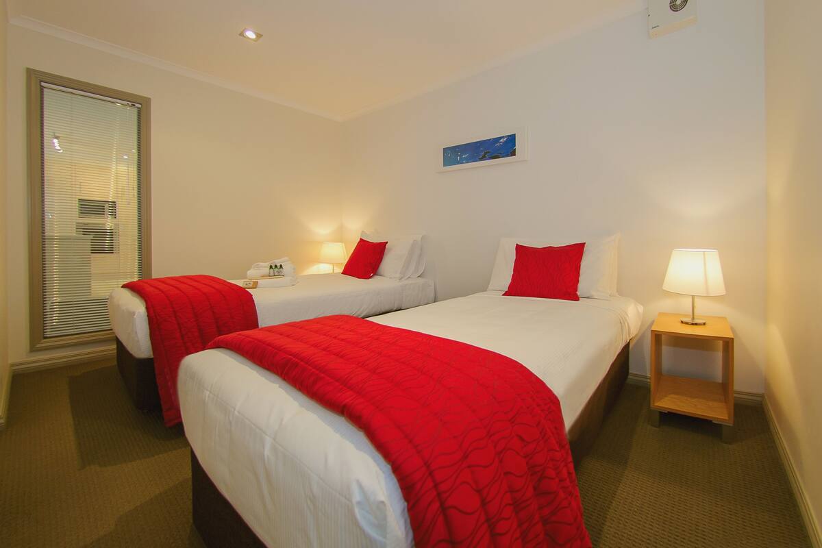 Habitación de hotel con dos camas, cada una con ropa de cama blanca y detalles rojos, una mesita de noche pequeña con lámpara y una ventana con persianas.