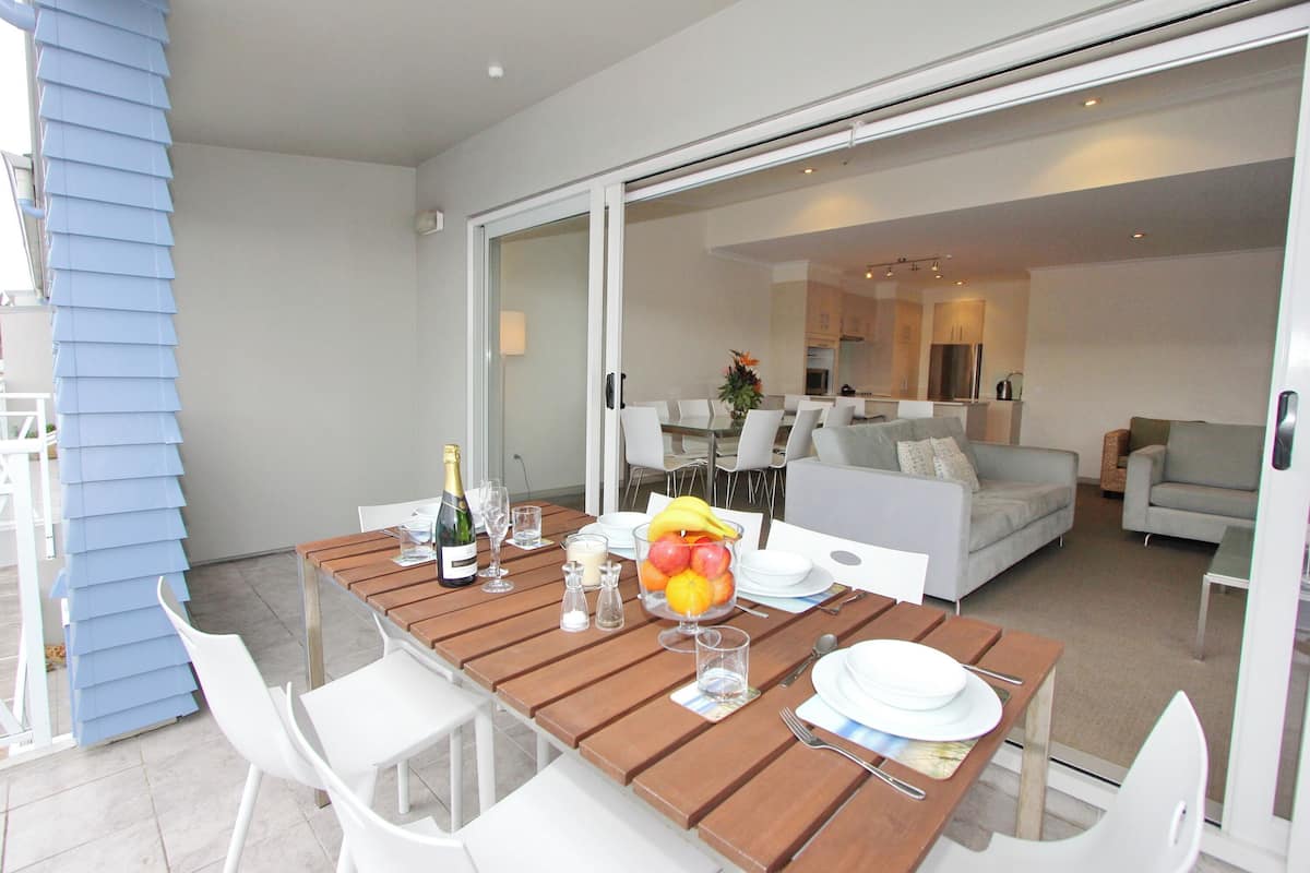 Un comedor con una mesa de madera preparada para cuatro personas, sillas blancas y un frutero.
