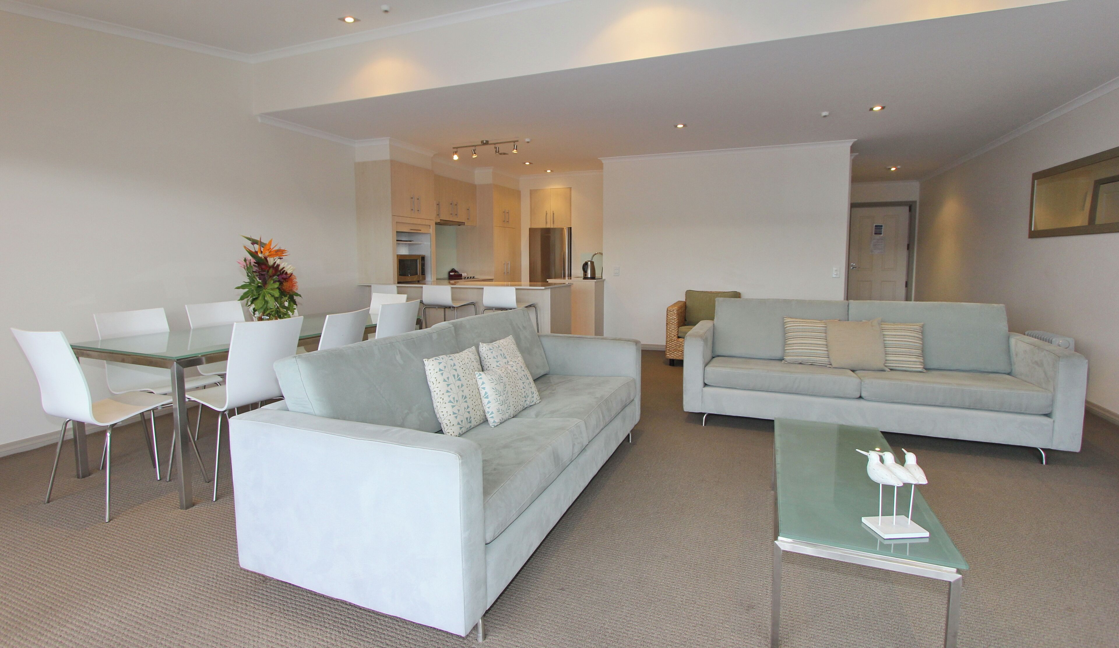 A modern living room with a grey sofa, a dining table with chairs, and a kitchen in the background.