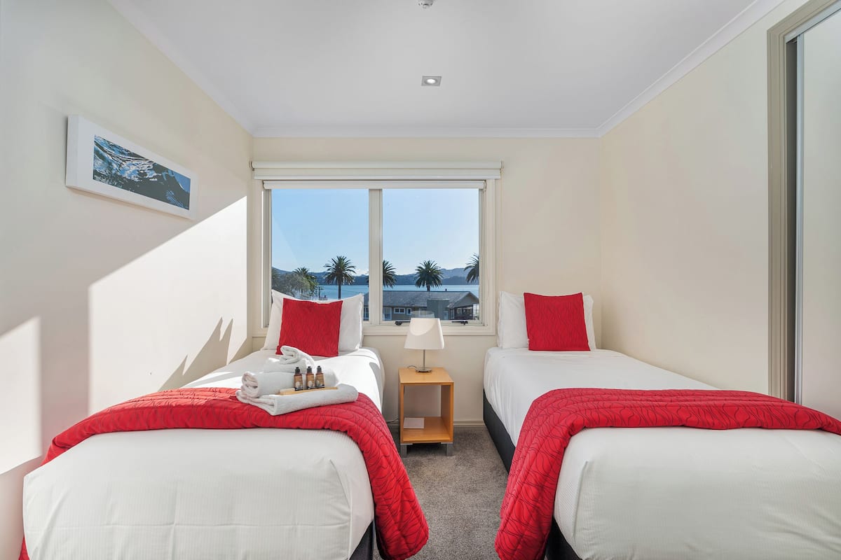 Una habitación de hotel con dos camas, una ventana que ofrece vistas a palmeras y un edificio, y una mesita con una lámpara y artículos de aseo.
