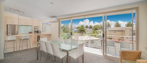 A modern kitchen with a dining area, large sliding glass doors leading to a balcony with a view of palm trees and buildings.