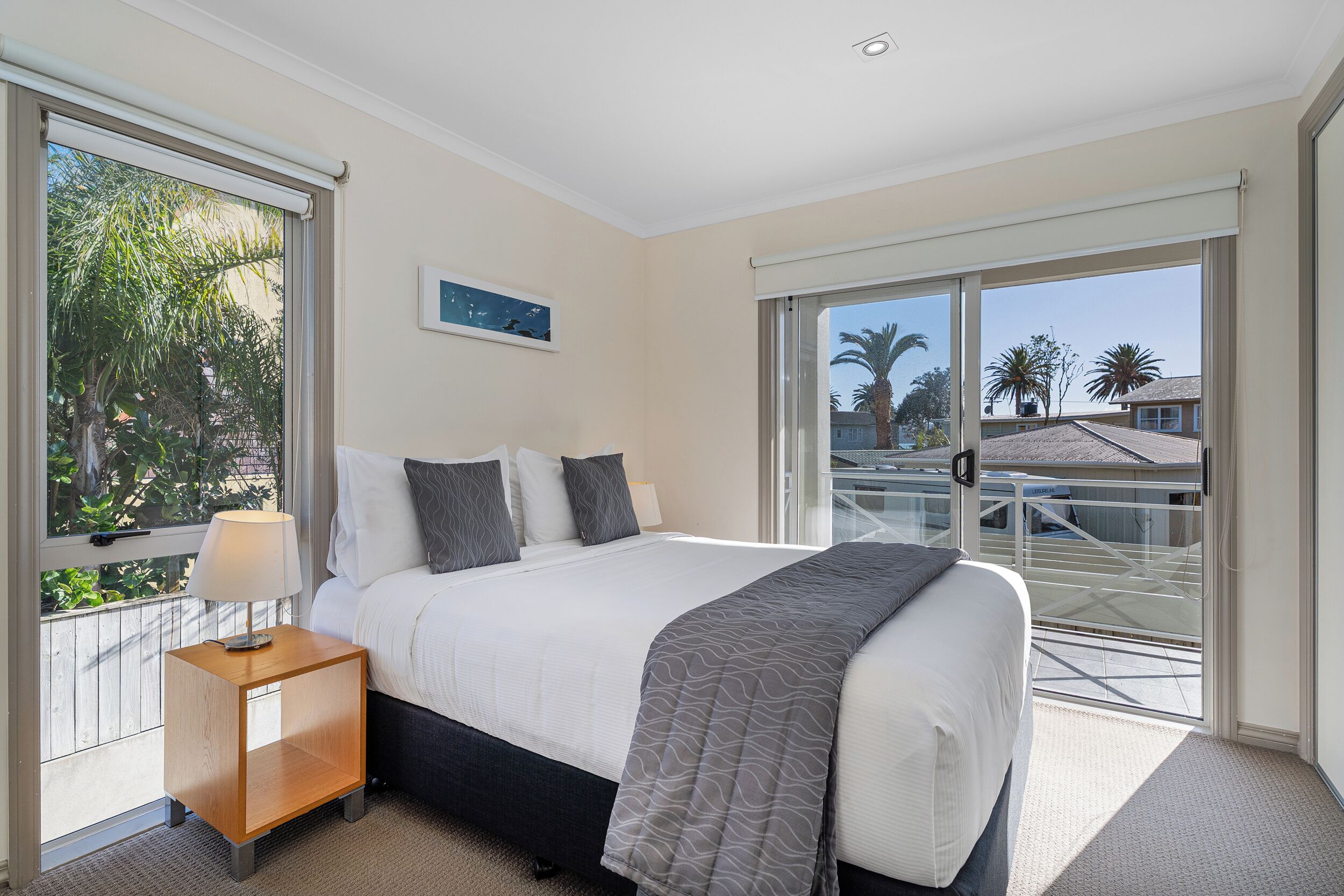 A bedroom with a large bed, a nightstand, a window with a view of palm trees, and a balcony with a glass railing.
