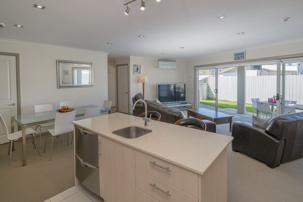 A modern kitchen with a central island, dining area with chairs, a sofa, and a TV. - Crowsnest Apartments (Whitianga)