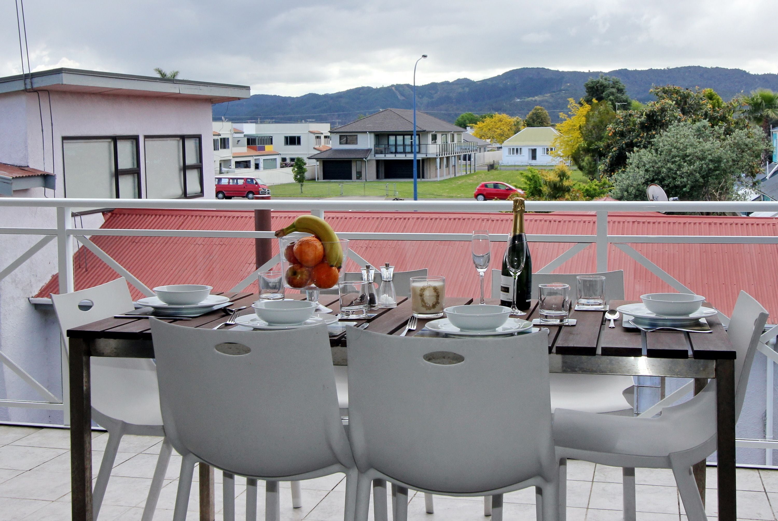 A dining table set with white chairs, plates, bowls, and a bottle on a balcony with a view of houses and mountains.