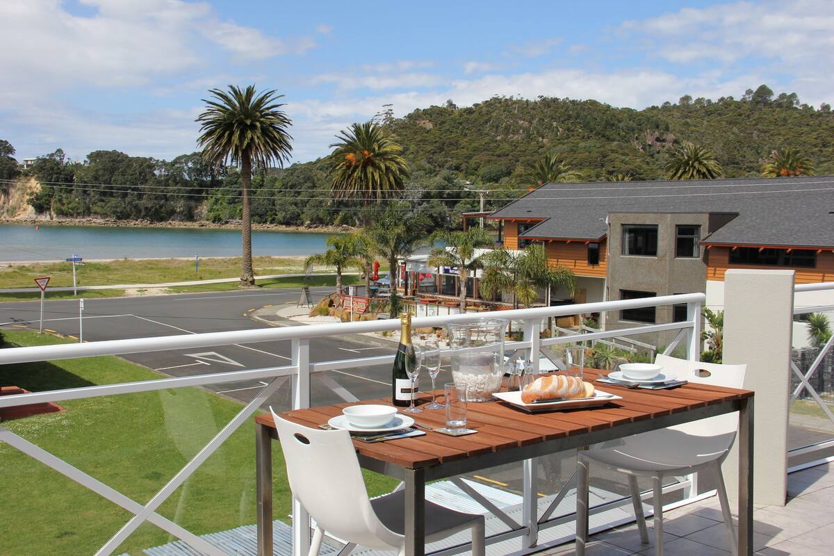 Un balcón con una mesa de madera lista para comer, con vistas panorámicas a una playa y una colina con árboles.