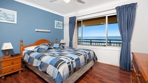 Ein Schlafzimmer mit Bett, Nachttisch, Lampe, Deckenventilator, einem Fenster mit Vorhängen und Blick aufs Meer.