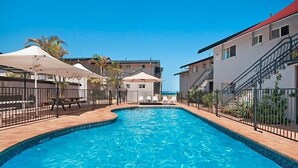 Ein Pool mit gewölbter Kante, umgeben von einem Zaun und Sonnenschirmen, mit Gebäuden im Hintergrund.