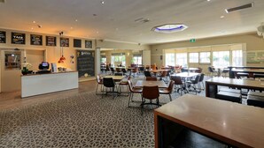 Una cafetería amplia con varias mesas y sillas, un área de recepción con mostrador y un monitor de computadora, y un menú en pizarra que anuncia las ofertas especiales.
