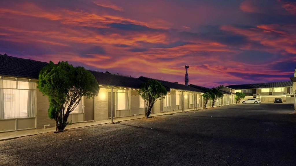 A row of motel-style buildings with individual rooms, a parking lot, and a clear sky.